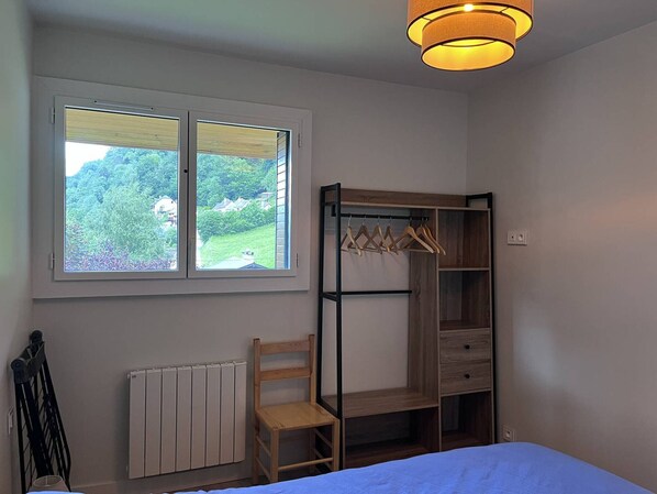 A bedroom with a bunk bed, a wooden chair, a radiator, a window with a view of greenery, and a hanging light fixture.