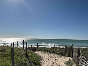 Un sentier de sable bordé d’une clôture en bois qui mène à l’océan, sous un ciel dégagé.