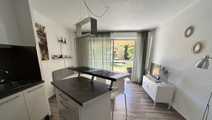A compact kitchen with a central island, a dining area with a table and chairs, a small white cabinet, and a view of the outside through a window.