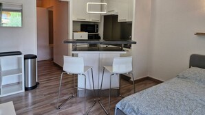A compact hotel room with a kitchenette, a bed, a small desk, and a trash bin.