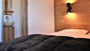 Une chambre à coucher avec une tête de lit en bois, un lit recouvert d’une couverture sombre, une table de chevet avec un miroir et un luminaire fixé au mur.
