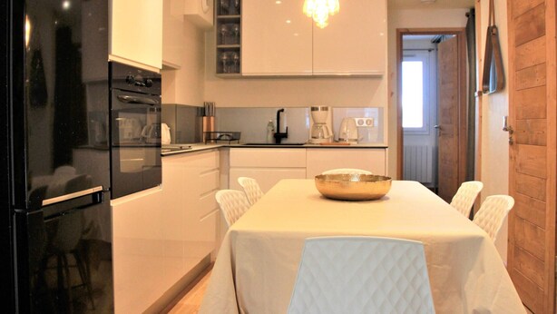 Une cuisine moderne avec des meubles blancs, un réfrigérateur noir, une table à manger recouverte d’une nappe blanche et un lustre.