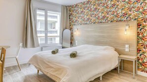 A bedroom with a bed, a chair, a window with curtains, a mirror, and a wall with a floral pattern. - Colivi Studio Tehuana Cosy Gare Annemasse (Annemasse)