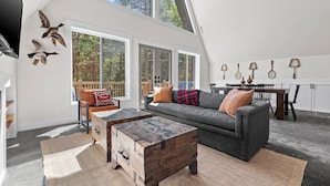 Ein geräumiger Loft mit hohen Decken, großen Fenstern, einer gemütlichen Sitzecke mit grauem Sofa und Holz-Couchtisch sowie einem Essbereich mit Holztisch und Stühlen.