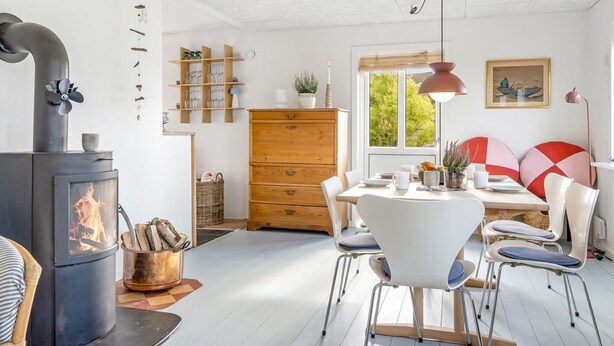 Una sala de estar acogedora con una estufa de leña, una zona de comedor con sillas blancas y un ventanal con vistas a la vegetación.