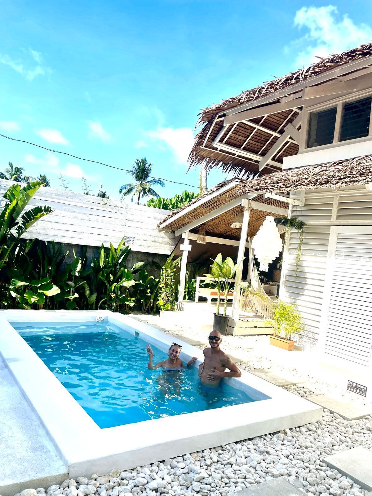 A swimming pool with two people, surrounded by a house with a thatched roof and a garden with tropical plants.