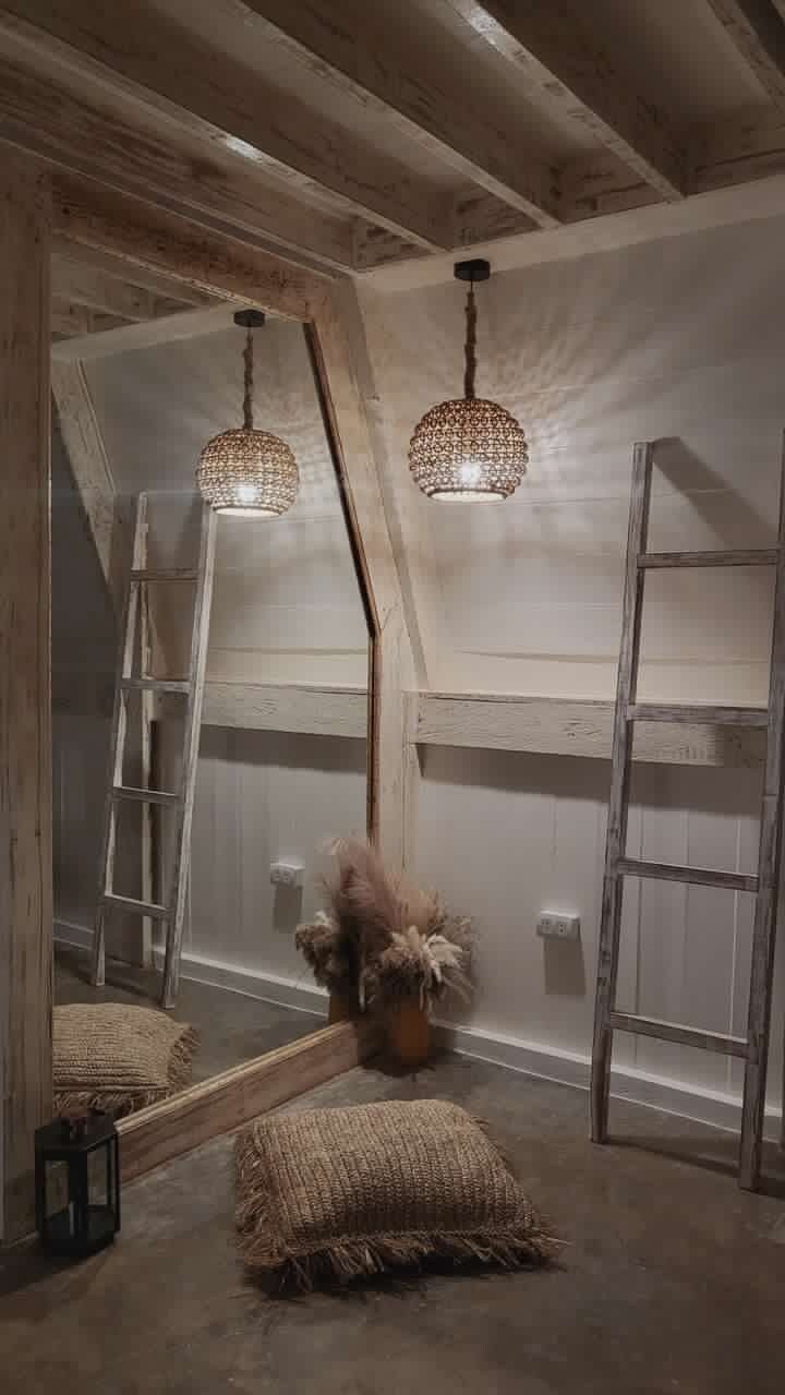 A rustic attic room with exposed wooden beams, a ladder, a hanging light fixture, and a large mirror.