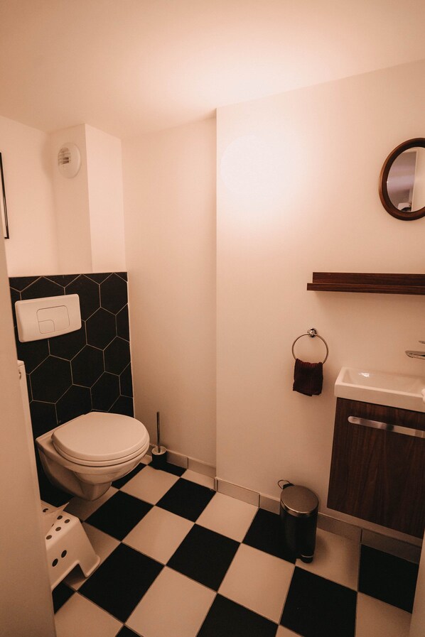 A small bathroom with a black and white checkered floor, a white toilet, a black hexagonal tile backsplash, a wooden shelf above the sink, and a round mirror on the wall. - L'Atelier de l'Horloger - 6 pers - 2 parkings - near train station (Besançon)