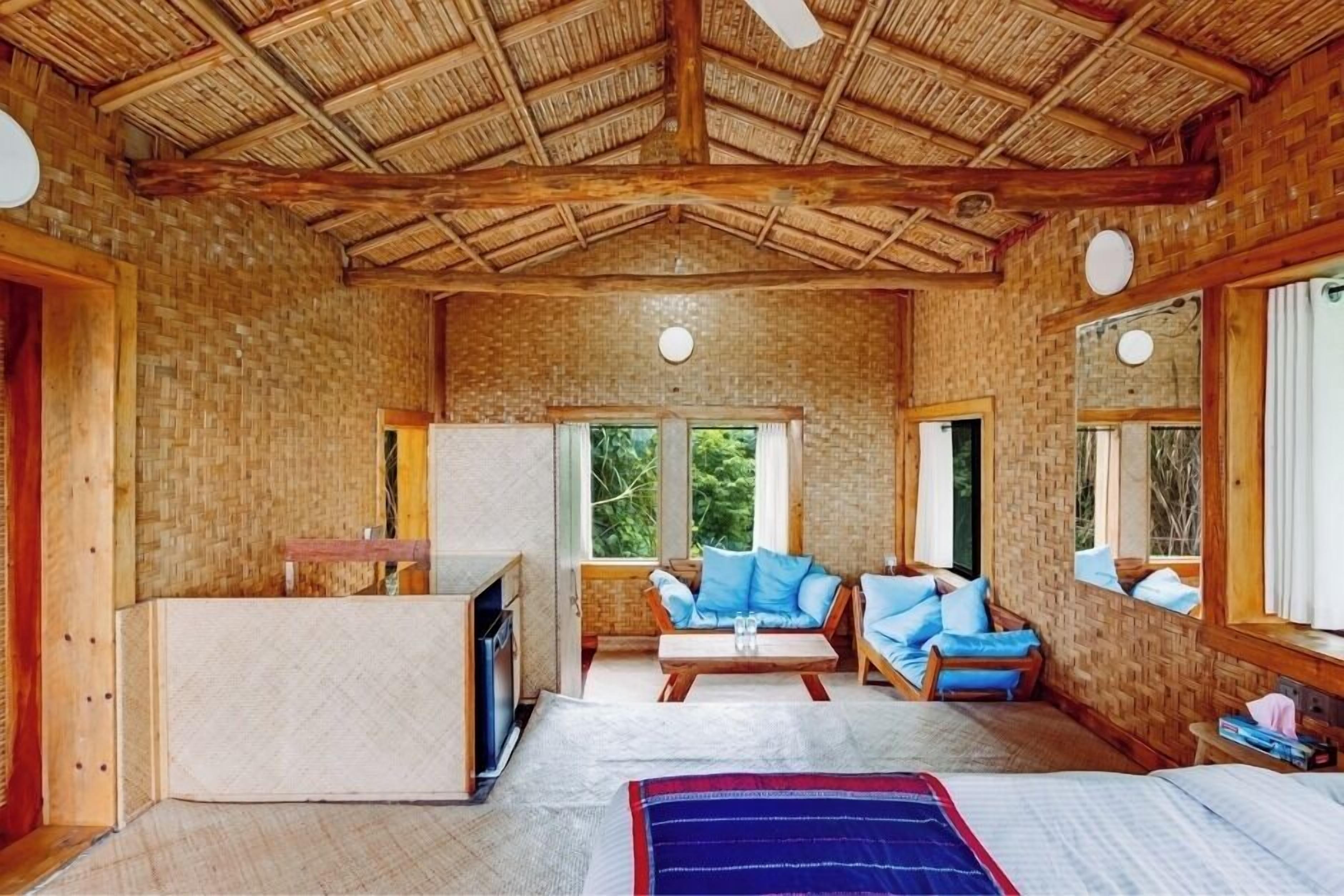 Habitación con techo de paja, paredes de ladrillo y ventanas amplias que dan a vistas de vegetación. Cuenta con una cama, un sofá y una mesita con un televisor.