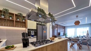 A modern kitchen with a central island, built-in oven, and stainless steel appliances. There are hanging plants, a wooden cutting board with fruits, and a knife block.
