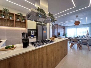 A modern kitchen with a central island, built-in oven, and stainless steel appliances. There are hanging plants, a wooden cutting board with fruits, and a knife block. - Modern and gorgeous beach condo (Punta Cana)