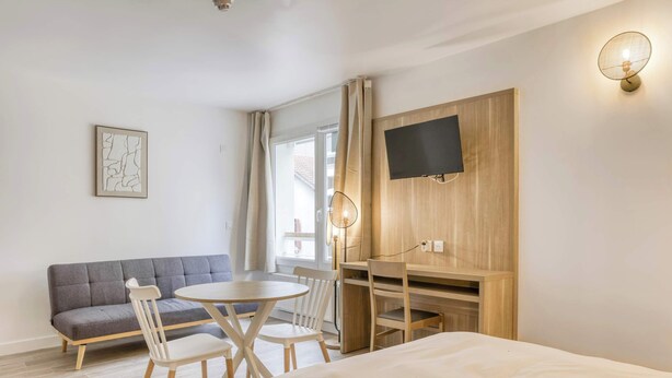 Ein modernes Hotelzimmer mit einem Bett, einer Couch, einem Esstisch mit Stühlen, einem an einer Holzplatte montierten Fernseher und einem Fenster mit Vorhängen.