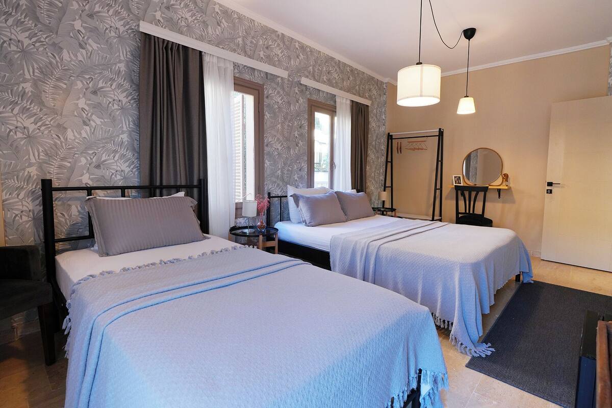 A hotel room with two beds, a small table, a mirror, and a window with curtains.
