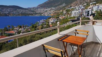 Uma varanda com mobiliário de madeira com vista para uma cidade costeira, com edifícios e uma montanha ao fundo.