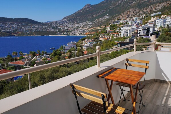 A balcony with wooden furniture overlooking a coastal town with buildings and a mountain in the background. - Kalkan Ayapa Hotel (Kaş)