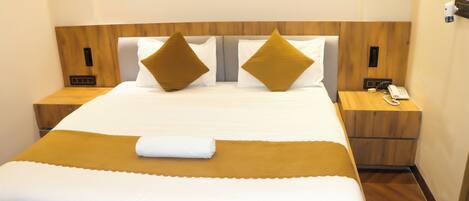 A neatly made bed with a white and mustard-colored bedspread, two mustard-colored pillows, and a white towel. There is a wooden headboard, a bedside table with a phone, and a wall-mounted light.