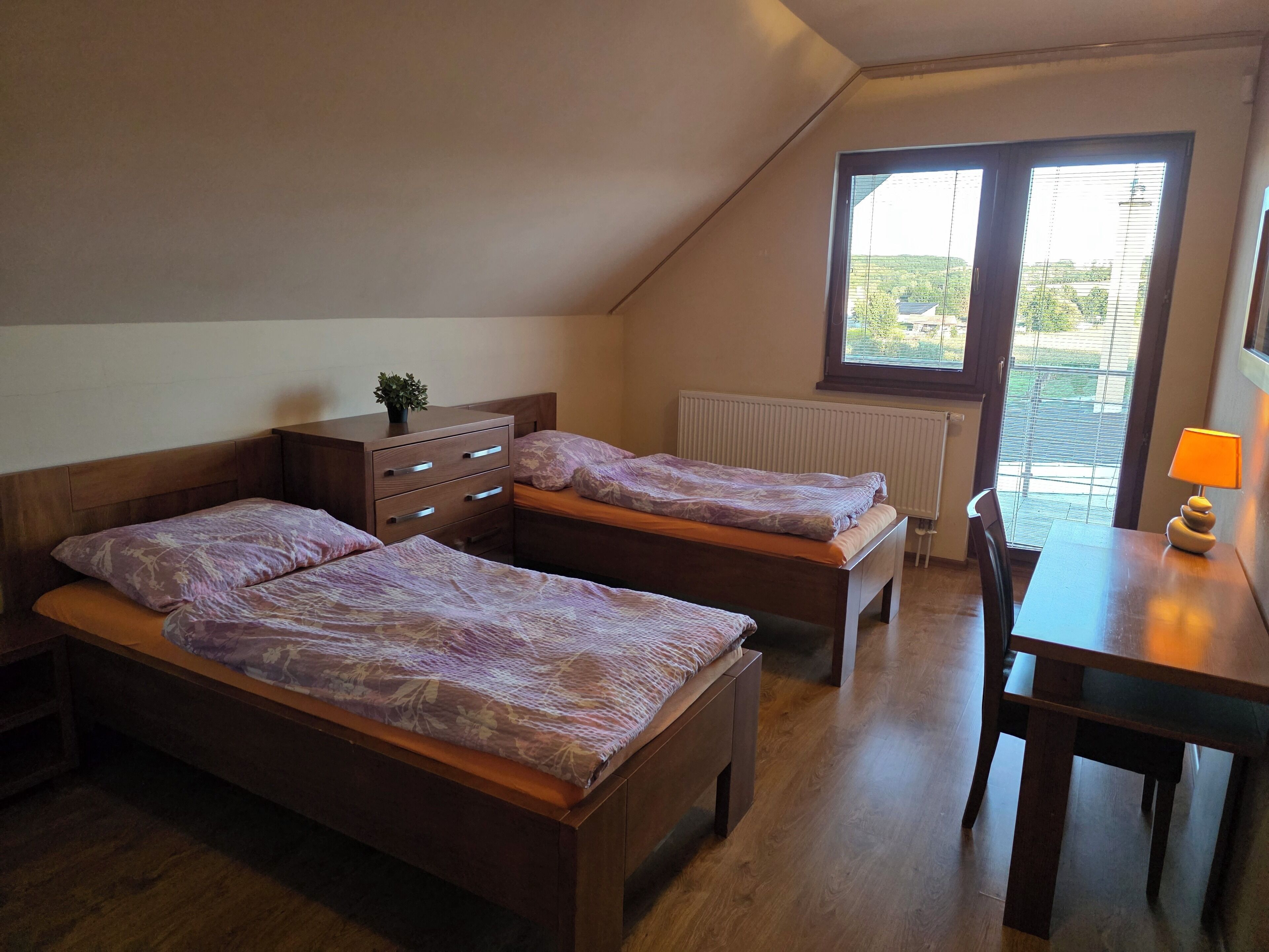 Ein Schlafzimmer mit zwei Holzbetten, einer Holzkommode, einem kleinen Tisch mit Lampe und einem Fenster mit Jalousien.