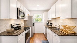 Una cocina estrecha con armarios blancos, un refrigerador de acero inoxidable, un microondas y una cafetera.