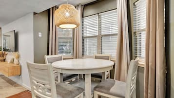 Un comedor con una mesa redonda, sillas blancas, una lámpara colgante de fibra y ventanas amplias con persianas.