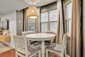 A dining area with a round table, white chairs, a woven pendant light, and large windows with blinds.