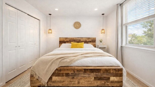 Un dormitorio con cabecera de madera, cama con ropa blanca y almohada amarilla, mesita de noche con lámpara y vista al exterior a través de una ventana.