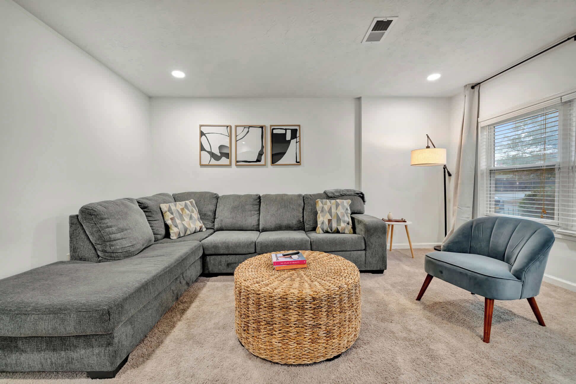 A modern living room with a grey sectional sofa, a round woven ottoman, and a blue armchair. There are three framed artworks on the wall, a floor lamp, and a window with blinds.