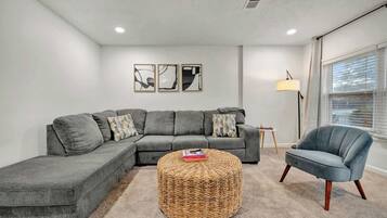 A modern living room with a grey sectional sofa, a round woven ottoman, and a blue armchair. There are three framed artworks on the wall, a floor lamp, and a window with blinds.
