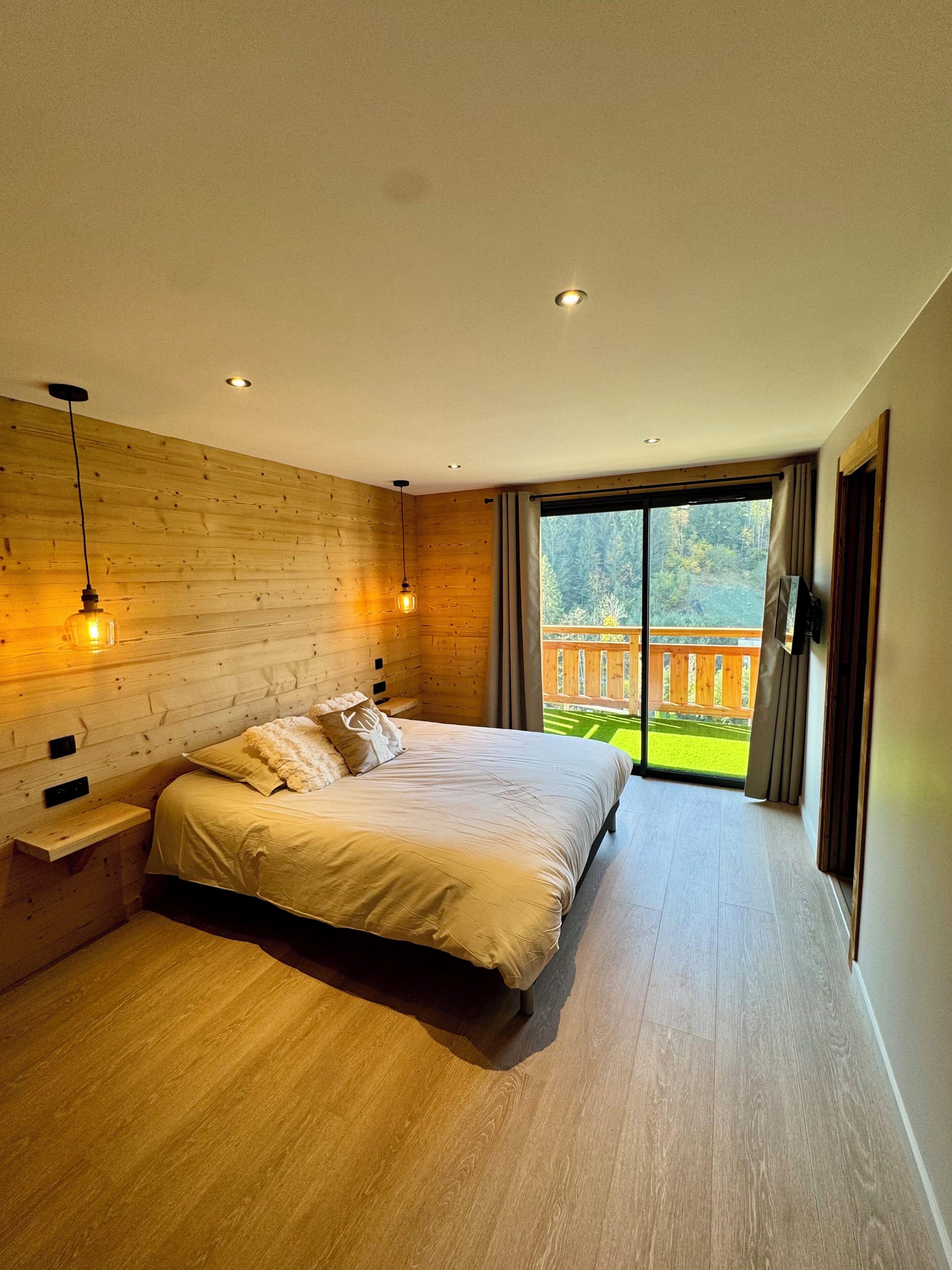 Een moderne slaapkamer met een groot bed, houten wanden en uitzicht op een houten terras en bomen.