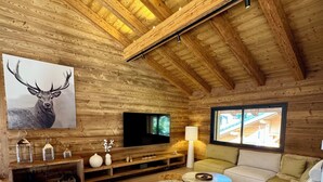 Ein Interieur einer Blockhütte mit Holzbalkendecke, einem aufgehängten Hirschgemälde, einem Fernseher, einer Couch und einem Fenster mit Blick auf ein Gebäude.