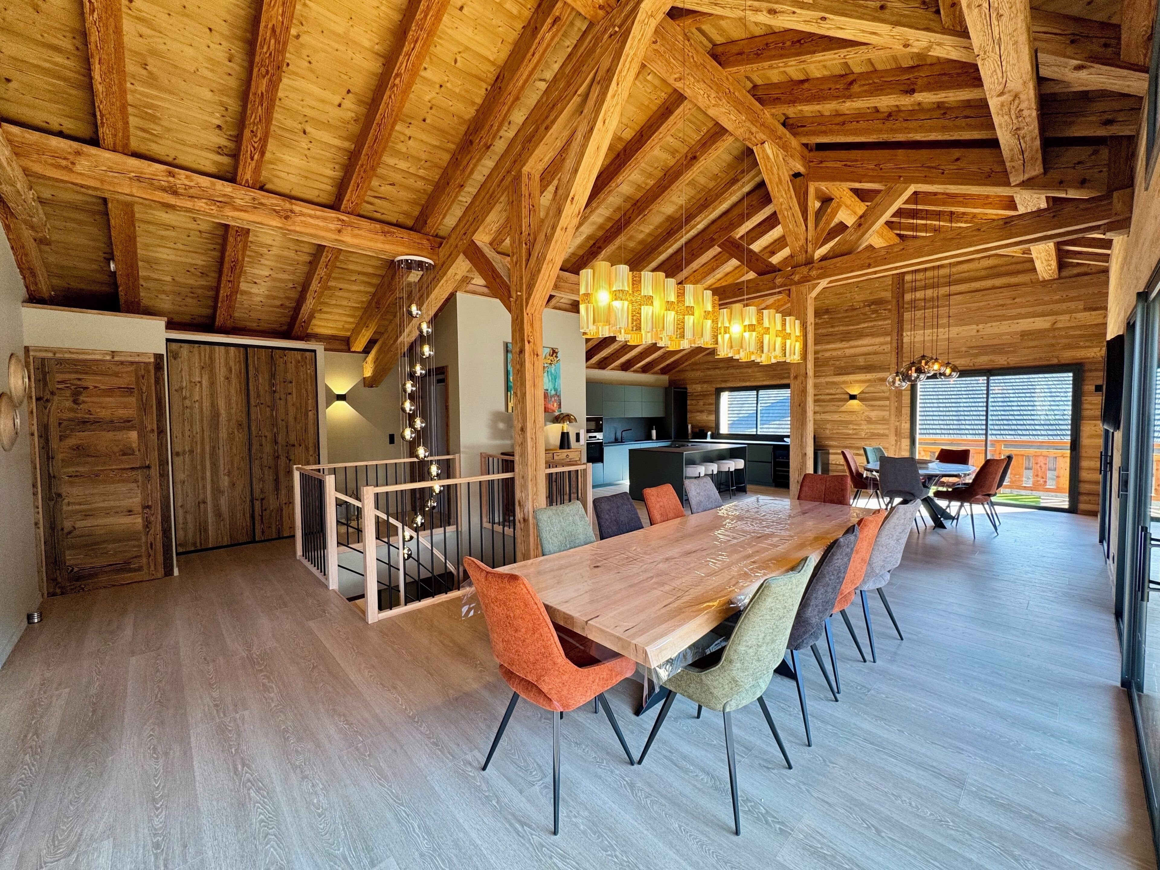 Un comedor espacioso con una larga mesa de madera y sillas coloridas, una cocina al fondo y una estructura de techo de madera.