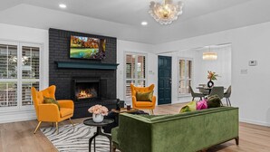 A living room with a fireplace, two armchairs, a sofa, a dining table with chairs, and a chandelier. - Bubble Bungalow - Cowboy Pool, Hot Tub & Firepit! (Fredericksburg)