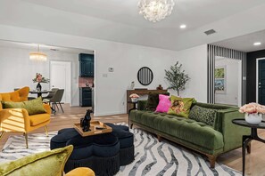 Un salon moderne avec un canapé vert, un fauteuil jaune et des poufs. On aperçoit un coin repas au fond et une cuisine à travers une porte ouverte.