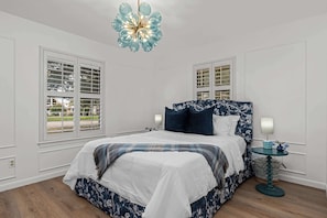 Ein Schlafzimmer mit einem Bett, zwei Fenstern, einer Kristallleuchter, einem Nachttisch mit Lampe und einem kleinen runden Tisch mit einer Vase.