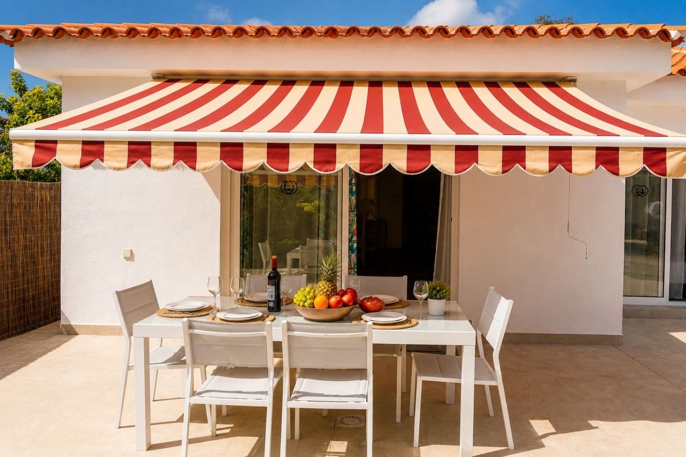 An outdoor dining area with a red and white striped awning, white chairs, a table set with plates, glasses, and a bowl of fruit.