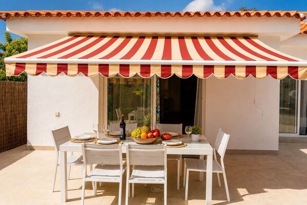An outdoor dining area with a red and white striped awning, white chairs, a table set with plates, glasses, and a bowl of fruit.