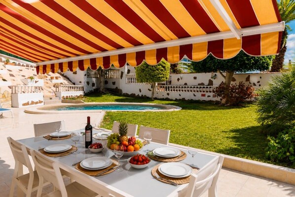 Un espace extérieur aménagé pour les repas, avec une piscine, des nappes blanches et un auvent à rayures.