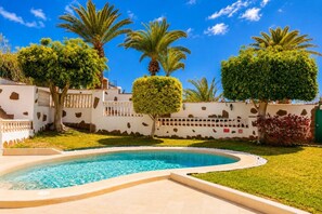 Une piscine à bordures courbes, entourée d’un mur blanc agrémenté d’éléments en pierre, et d’une végétation luxuriante comprenant des palmiers et un arbuste taillé avec soin.