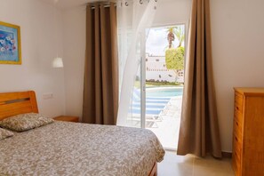 A bedroom with a bed, a wooden dresser, a painting on the wall, and a view of a pool and palm trees through a glass door.