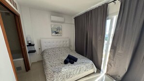 Une chambre à coucher comprenant un lit, une tête de lit, une table de chevet, un climatiseur fixé au mur et une fenêtre avec des rideaux.