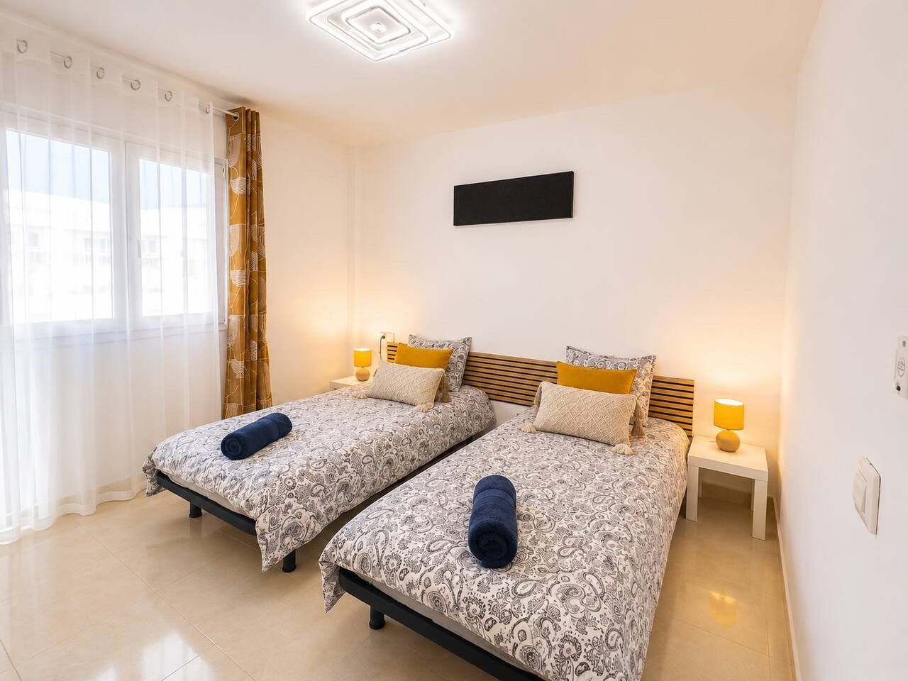 Habitación de hotel con dos camas, cada una con colcha y almohadas estampadas, una mesita de noche con lámpara y un ventanal con cortinas traslúcidas.