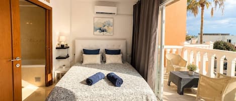 Un dormitorio con cama, mesita de noche, ventana y balcón con mobiliario exterior.