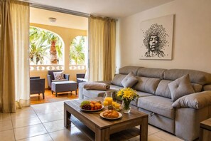 Una sala de estar con un sofá gris, una mesa de centro con frutas y un vaso de jugo de naranja, y un cuadro en la pared.