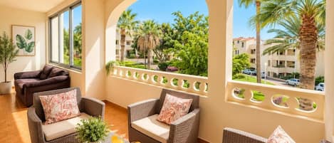 A sunlit lounge area with wicker furniture, a fruit tray, and a view of palm trees and buildings.