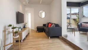 Un salon moderne avec un canapé, une table basse et un téléviseur fixé au mur. Un miroir rond est accroché au mur, et une petite table d’appoint avec une plante se trouve à proximité.