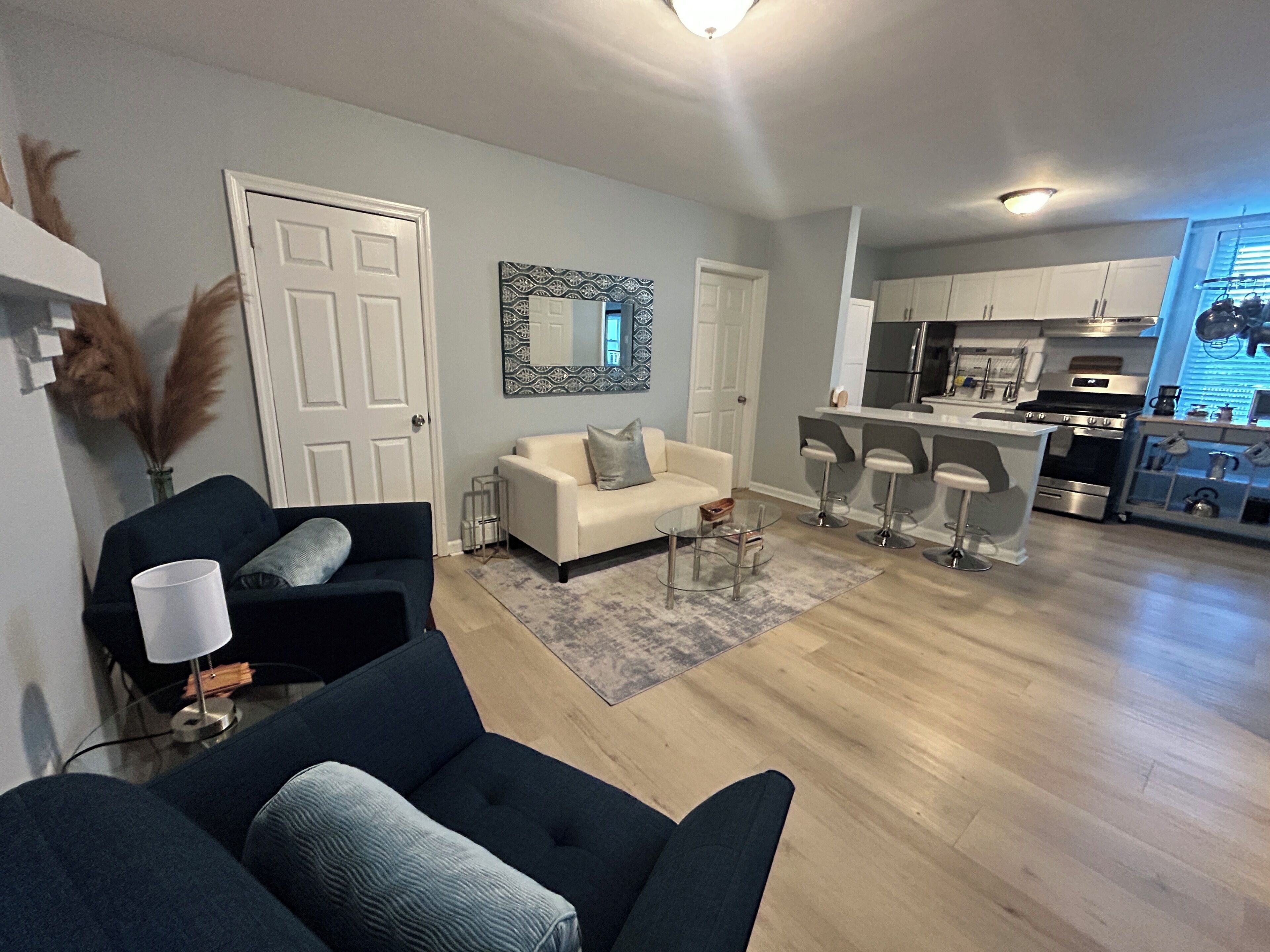 A living room with a sofa, a coffee table, and a TV. There is a kitchen area with a refrigerator, stove, and bar stools.