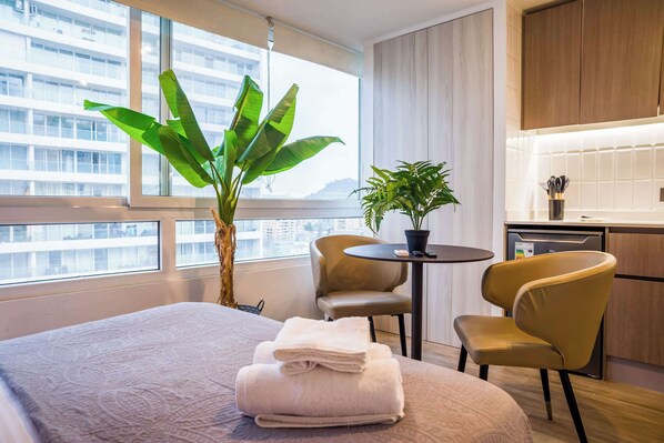 A hotel room with a bed, a small table with chairs, a potted plant, and a view of a cityscape through large windows.