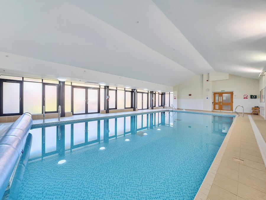 A large indoor swimming pool with clear glass walls, a tiled floor, and a wooden door with emergency exit signs.