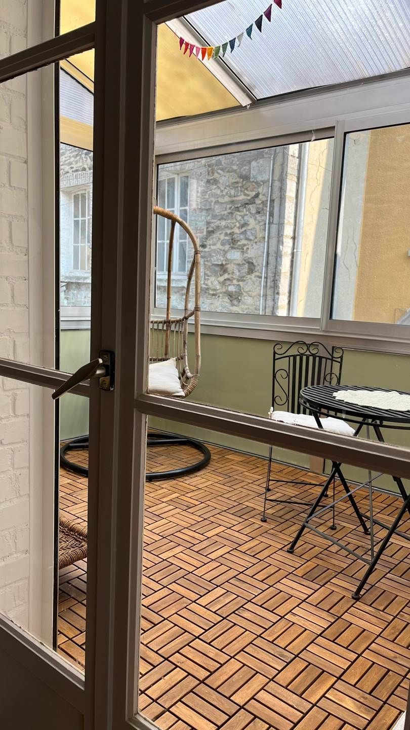 Une porte ouverte donne sur une pièce d’où l’on aperçoit un mur de briques, une chaise en osier, une table avec un coussin et un sol à motifs.