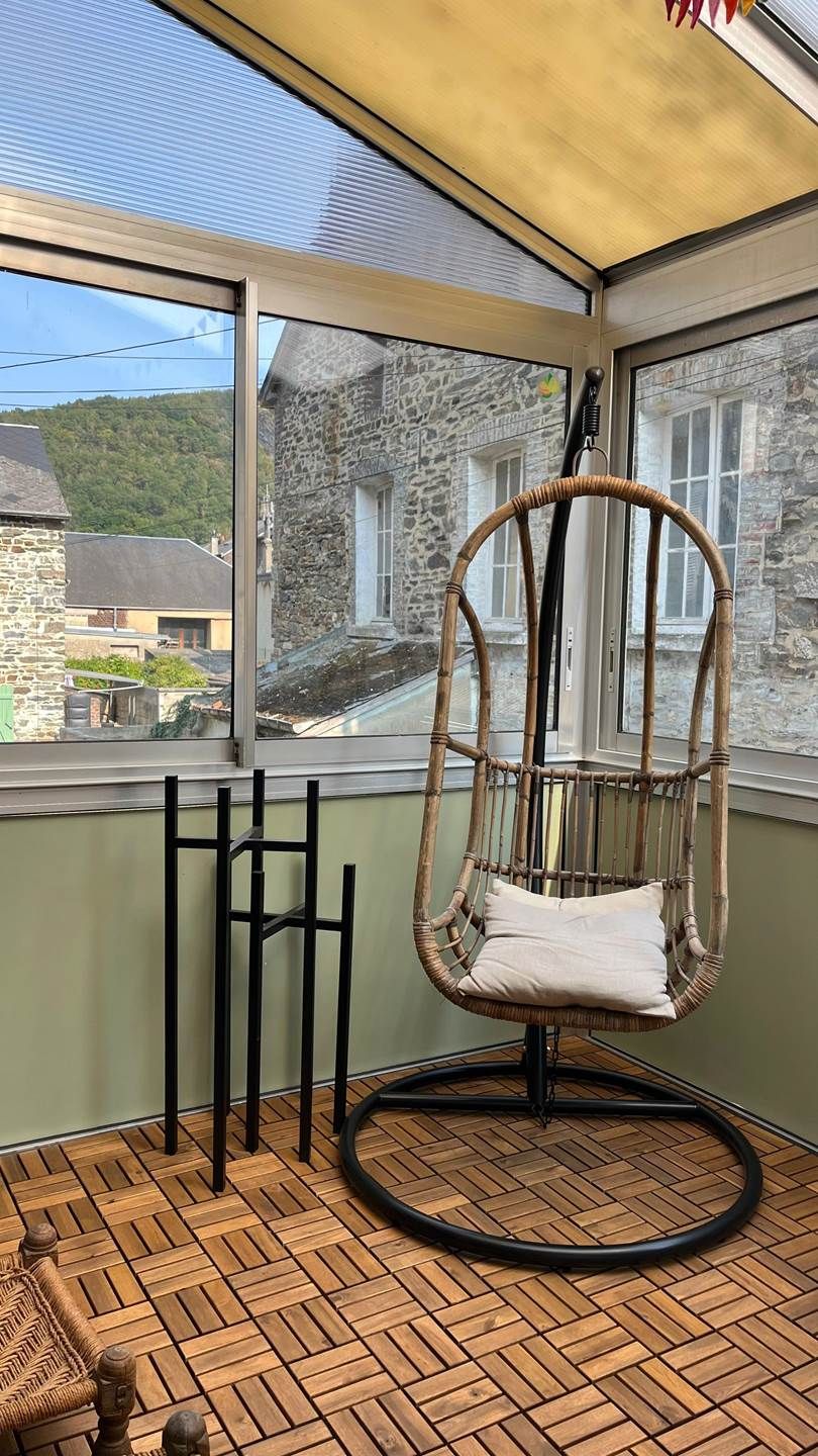 Une chaise en osier avec un coussin, dans une pièce dont le sol est recouvert d’un parquet à motifs en chevrons, avec de grandes fenêtres et une vue sur un bâtiment en pierre.