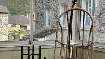 Une chaise en osier avec un coussin, dans une pièce dont le sol est recouvert d’un parquet à motifs en chevrons, avec de grandes fenêtres et une vue sur un bâtiment en pierre.